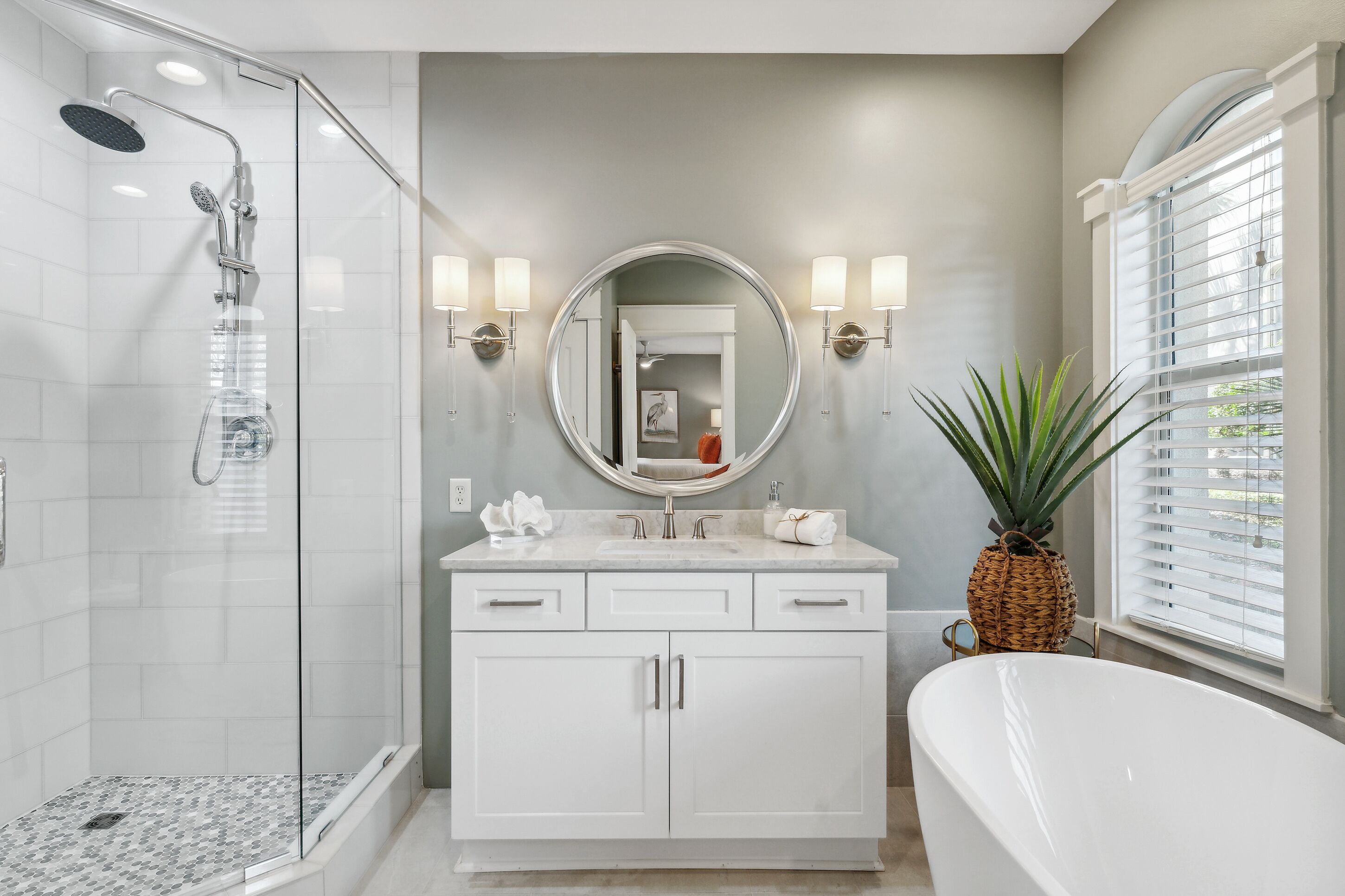 A modern bathroom with a glass-enclosed shower, a freestanding bathtub, and a vanity with a round mirror and wall-mounted lights.