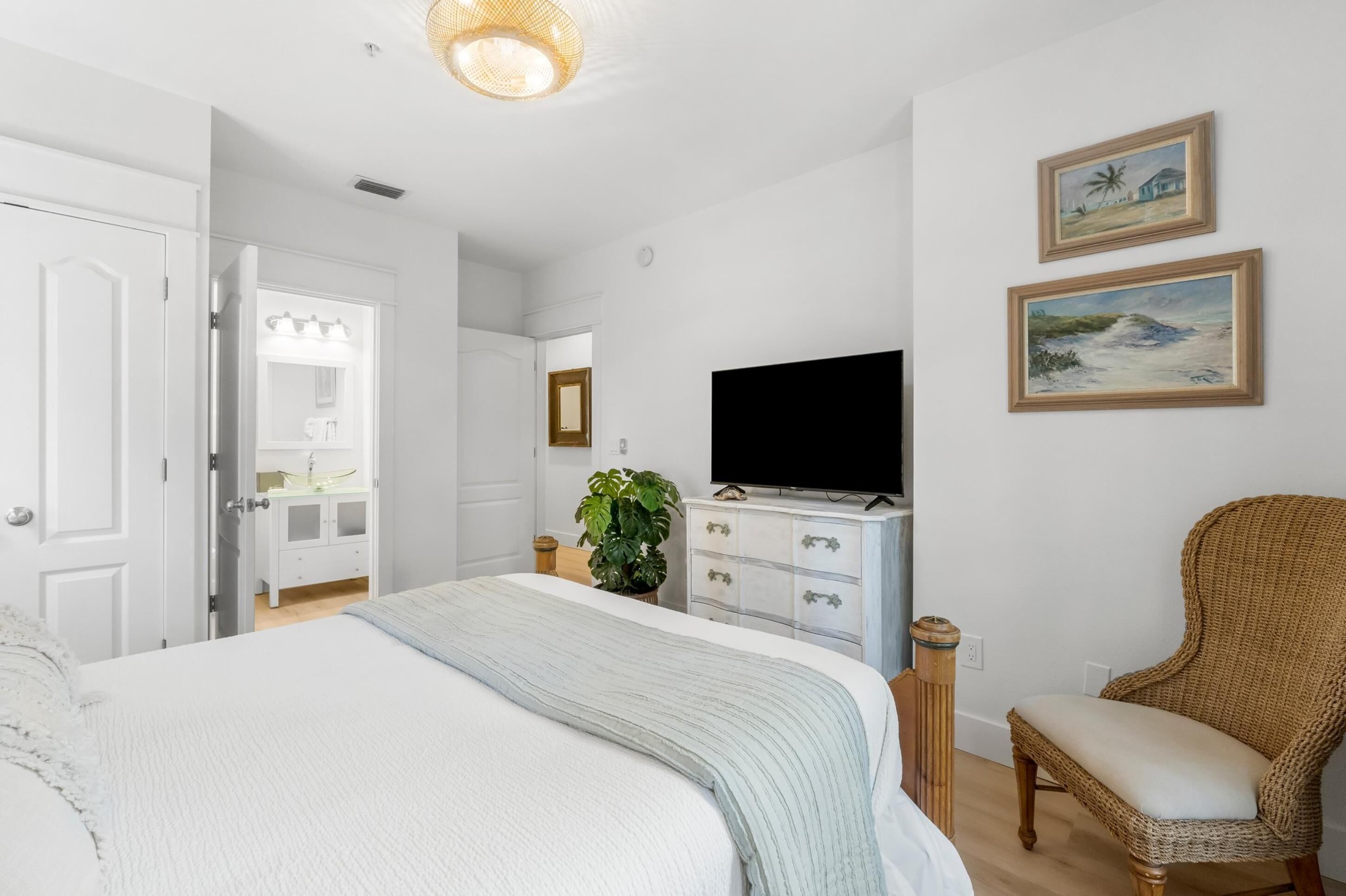 Une chambre à coucher comprenant un lit, un téléviseur, un fauteuil en osier et deux tableaux encadrés accrochés au mur.