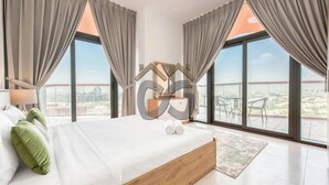 A modern bedroom with a large bed, two windows with curtains, a view of the city, and a balcony with chairs. - Serene 2 Bedroom Apartment in Dubai Silicon Oasis (دبي)