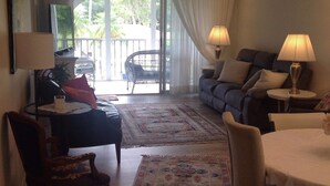 A living room with a sofa, a chair, a small table, and a lamp. There is a large window with curtains, and a view of greenery outside. - It is at the heart of Naples. Minutes from beach. Condo w/ pools, tennis, golf.  (Naples)