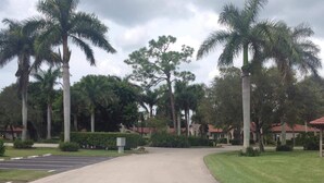 A paved driveway leading to a building with a red roof, surrounded by palm trees and greenery. - It is at the heart of Naples. Minutes from beach. Condo w/ pools, tennis, golf.  (Naples)