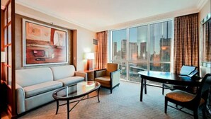Habitación de hotel con sofá, sillón, mesa de centro y escritorio con una computadora portátil. Un ventanal grande ofrece vista a la ciudad.