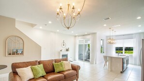 A spacious living room with a brown leather sofa, a glass coffee table, and a chandelier. There is a kitchen area with a bar and large windows. - Stunning Brand New 3 Bedroom, 2.5 Bathroom Luxury Townhome with Loft. (Deerfield Beach)