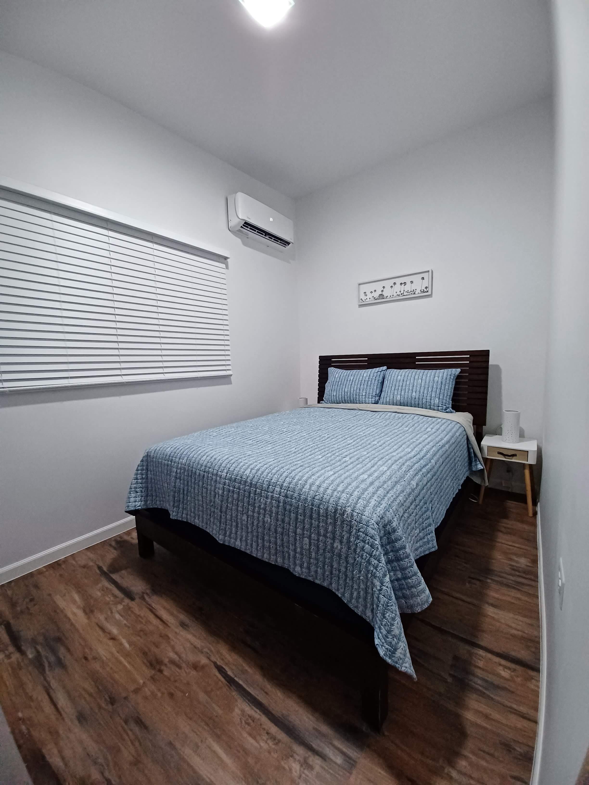 Une chambre à coucher comprenant un lit, un climatiseur, une fenêtre avec des stores et une petite table de chevet avec une lampe.