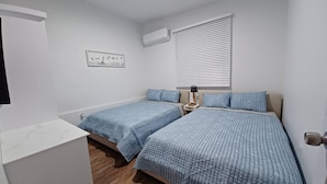 Une chambre avec deux lits, une fenêtre avec des stores, un appareil de climatisation et une petite table de chevet avec une lampe.