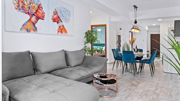 Ein modernes Wohnzimmer mit einem grauen Ecksofa, einem Essbereich mit blauen Stühlen und einer Küche im Hintergrund.