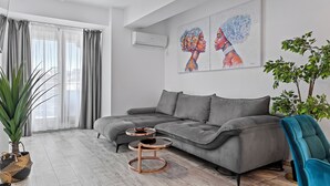 Ein modernes Wohnzimmer mit einem grauen Ecksofa, zwei runden Couchtischen, einer großen Topfpflanze und einem türkisfarbenen Sessel.