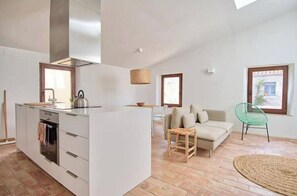 Eine moderne Küche mit Kochinsel, ein Essbereich mit Sofa und Stühlen sowie ein Blick ins Freie durch ein Fenster.