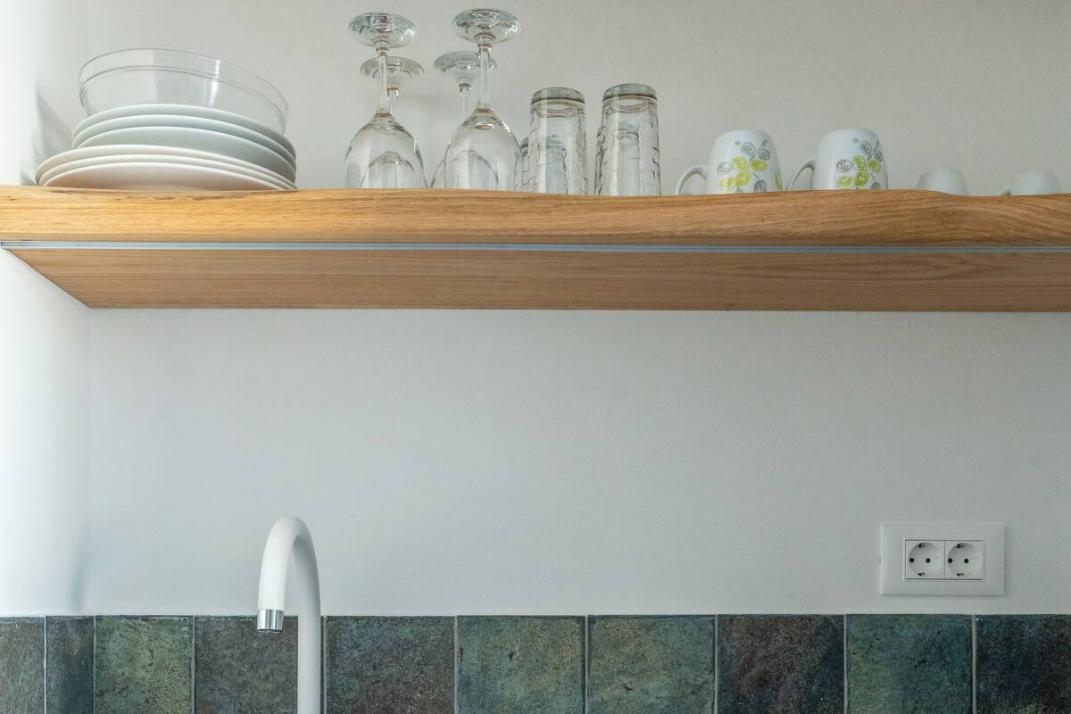 Ett kök med en vit diskho, gröna kakelplattor ovanför diskbänken, hyllor i trä och en vit diskmaskin.