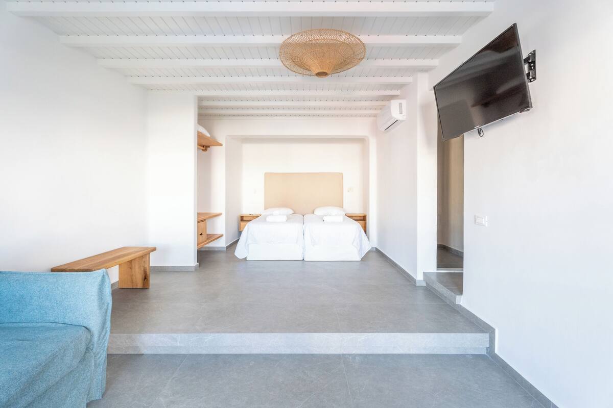 Ett modernt hotellrum med en stor säng, en tv som är monterad på väggen, en träbänk och en vävd taklampa.