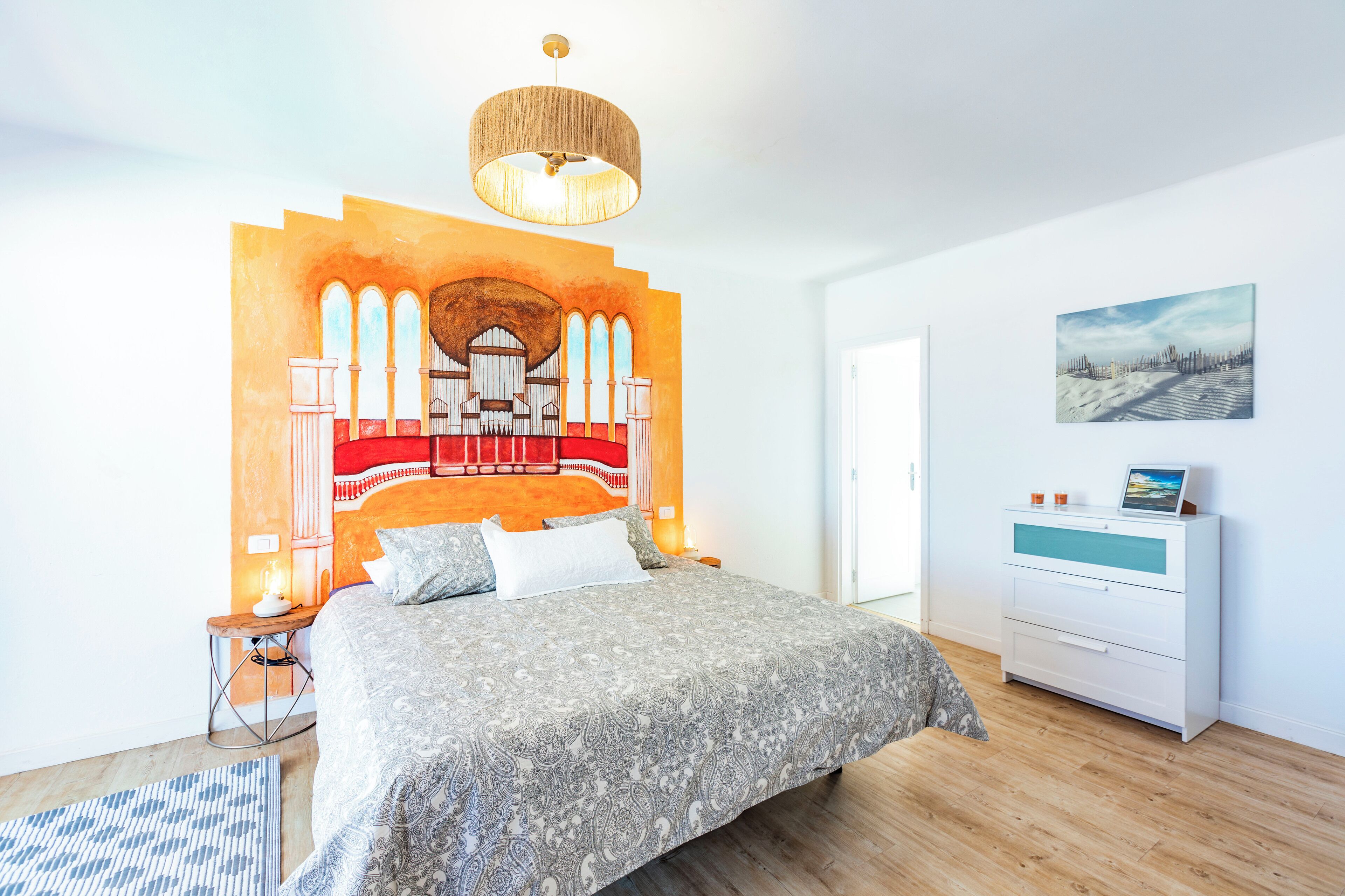 A modern bedroom with a large bed, a wall mural, a wooden floor, a white dresser, and a hanging light fixture.