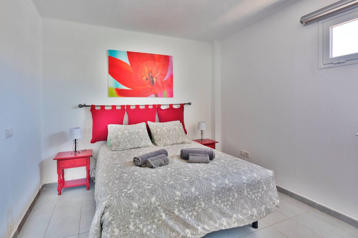 Quarto com cama, mesas de cabeceira vermelhas, cabeceira vermelha e um quadro floral grande na parede.