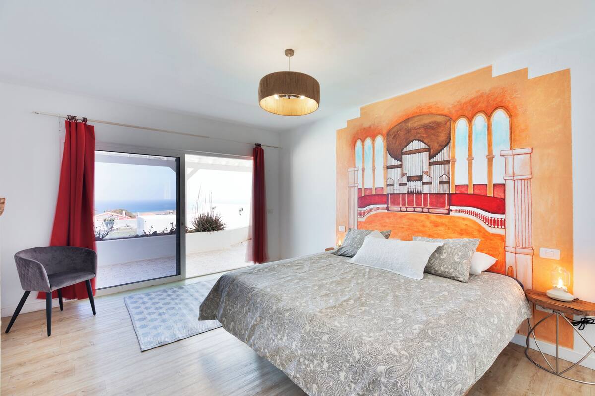 Quarto com cama grande, uma cadeira, um quadro e vista para o exterior.