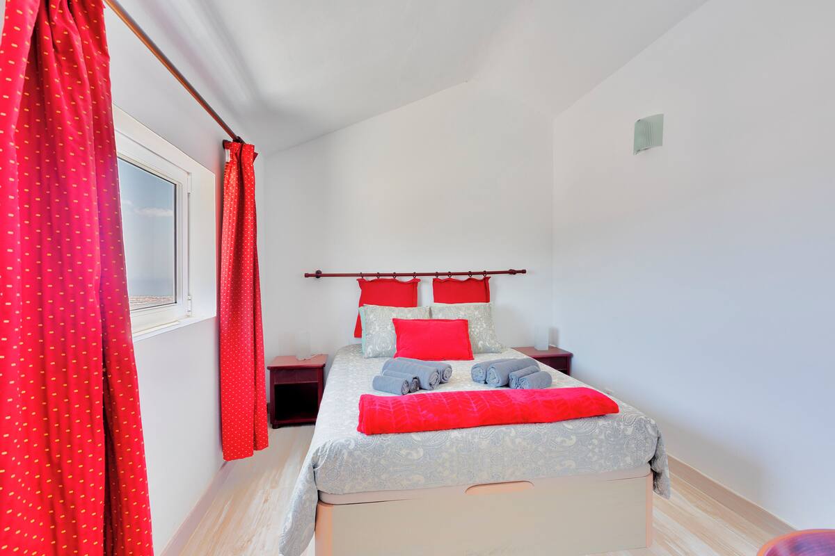 Cama bem arrumada com roupa de cama vermelha e cinza, uma janela com cortinas vermelhas e uma mesa de cabeceira com abajur.