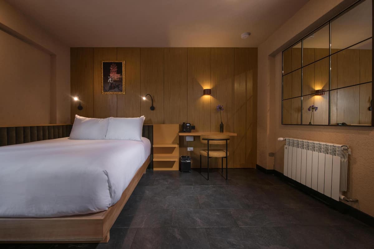 A hotel room with a bed, a small table, a chair, a radiator, and a wall-mounted mirror.