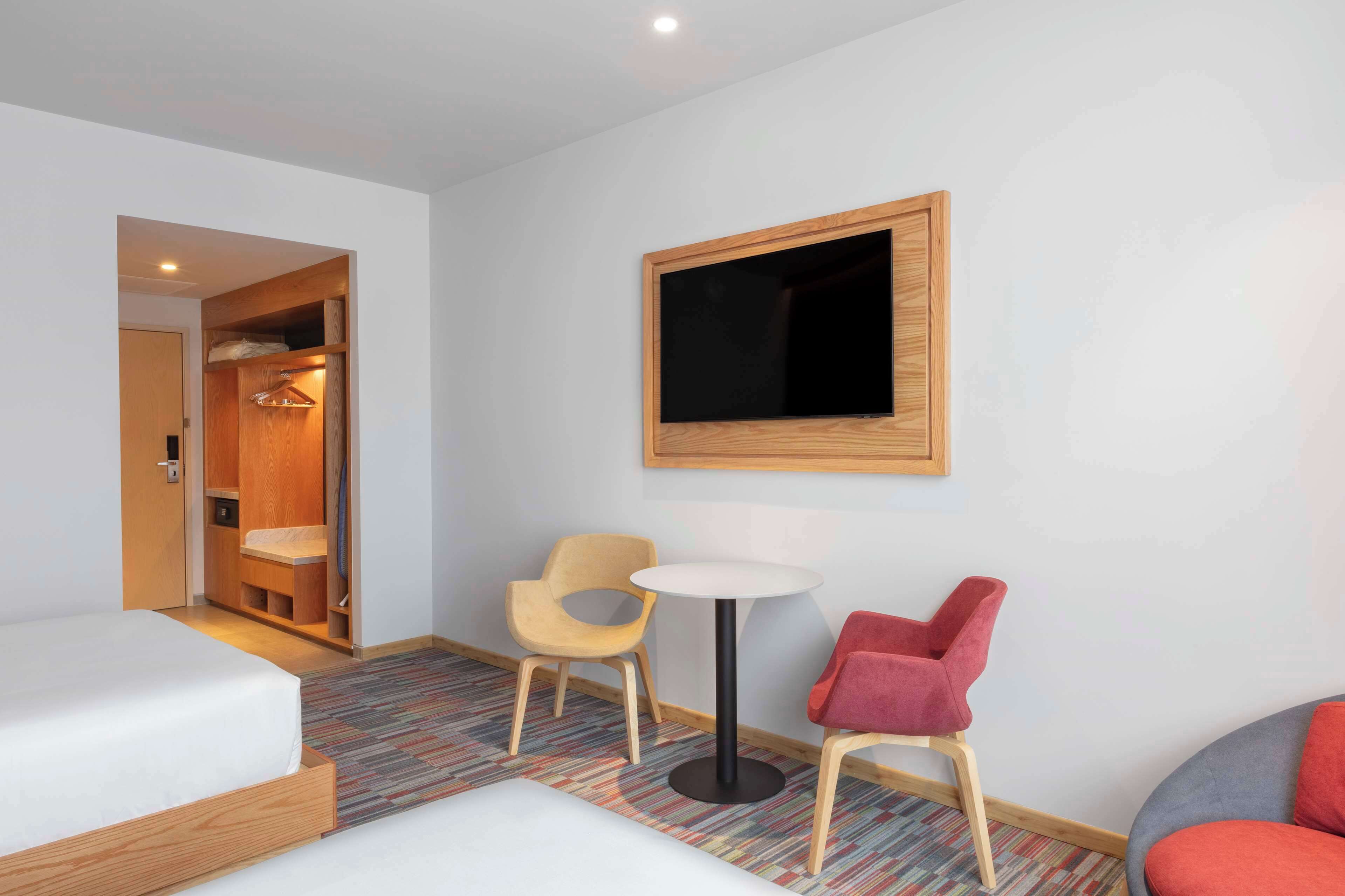 A hotel room with a bed, a small round table, two chairs, a wardrobe, and a wall-mounted TV.