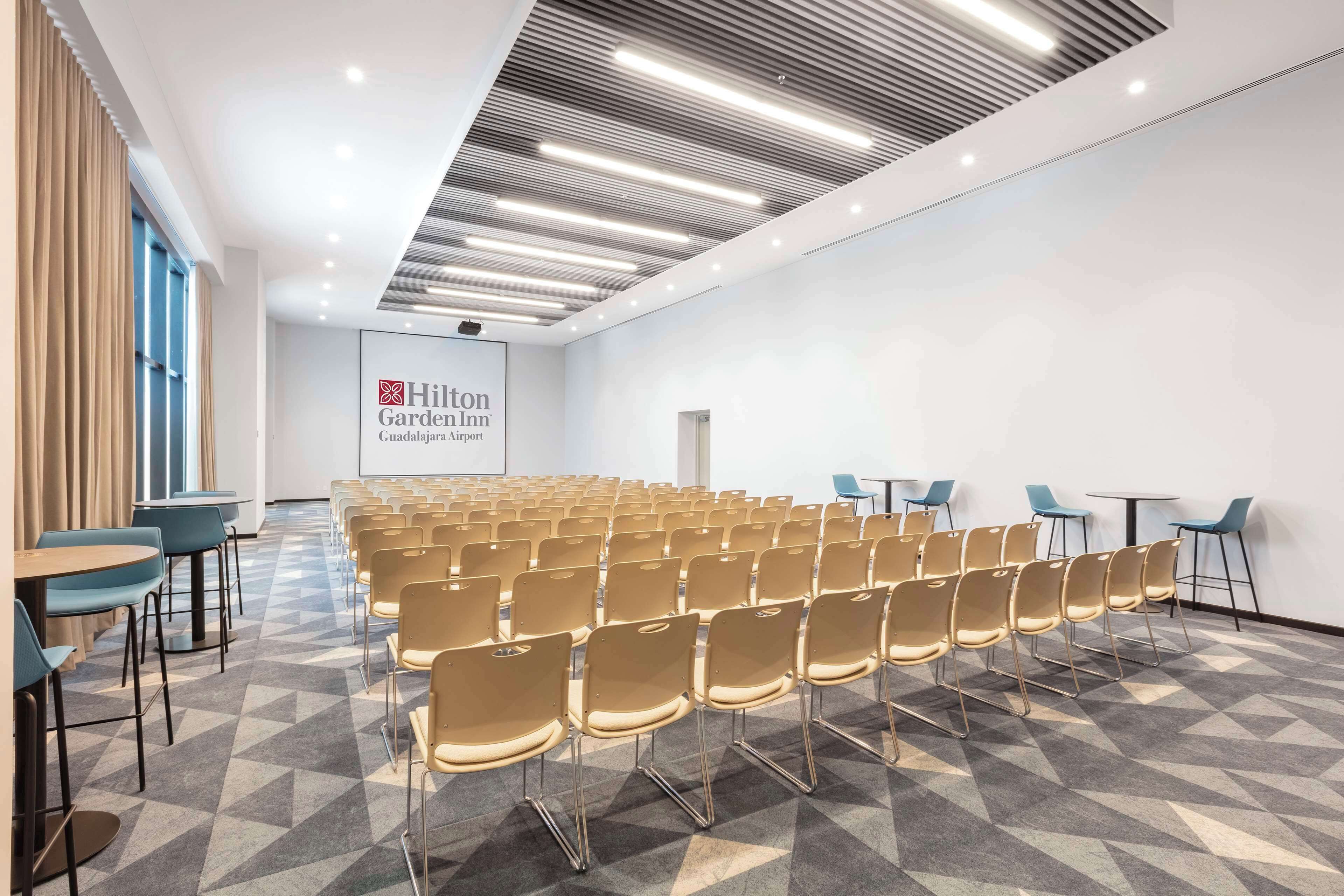 A conference room with rows of chairs, a projector screen, and a sign for Hilton Garden Inn Guadalajara Airport.