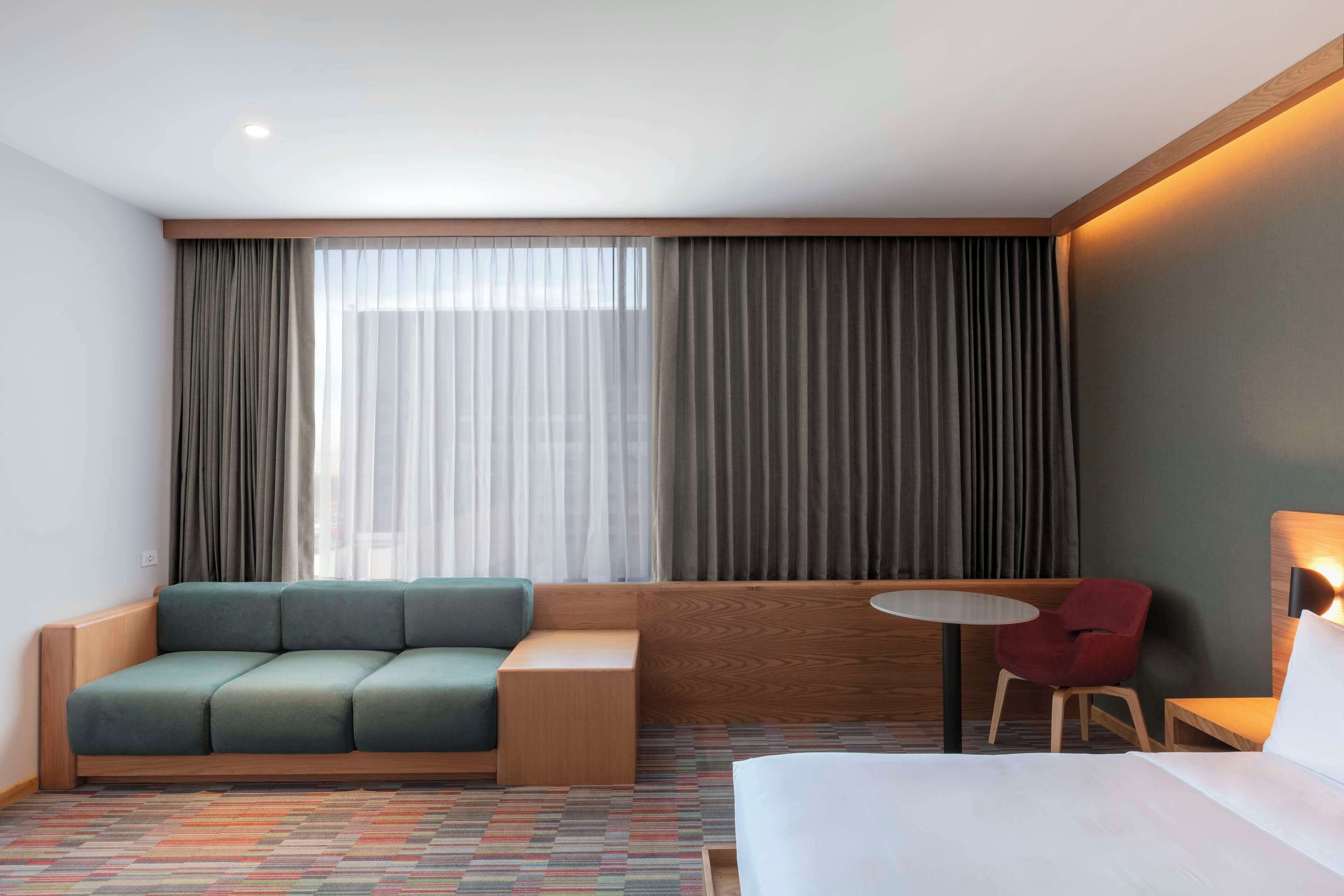 Una habitación de hotel con una cama, un sofá verde, una silla roja, una mesita pequeña y un ventanal grande con cortinas.