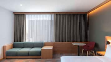 Una habitación de hotel con una cama, un sofá verde, una silla roja, una mesita pequeña y un ventanal grande con cortinas.