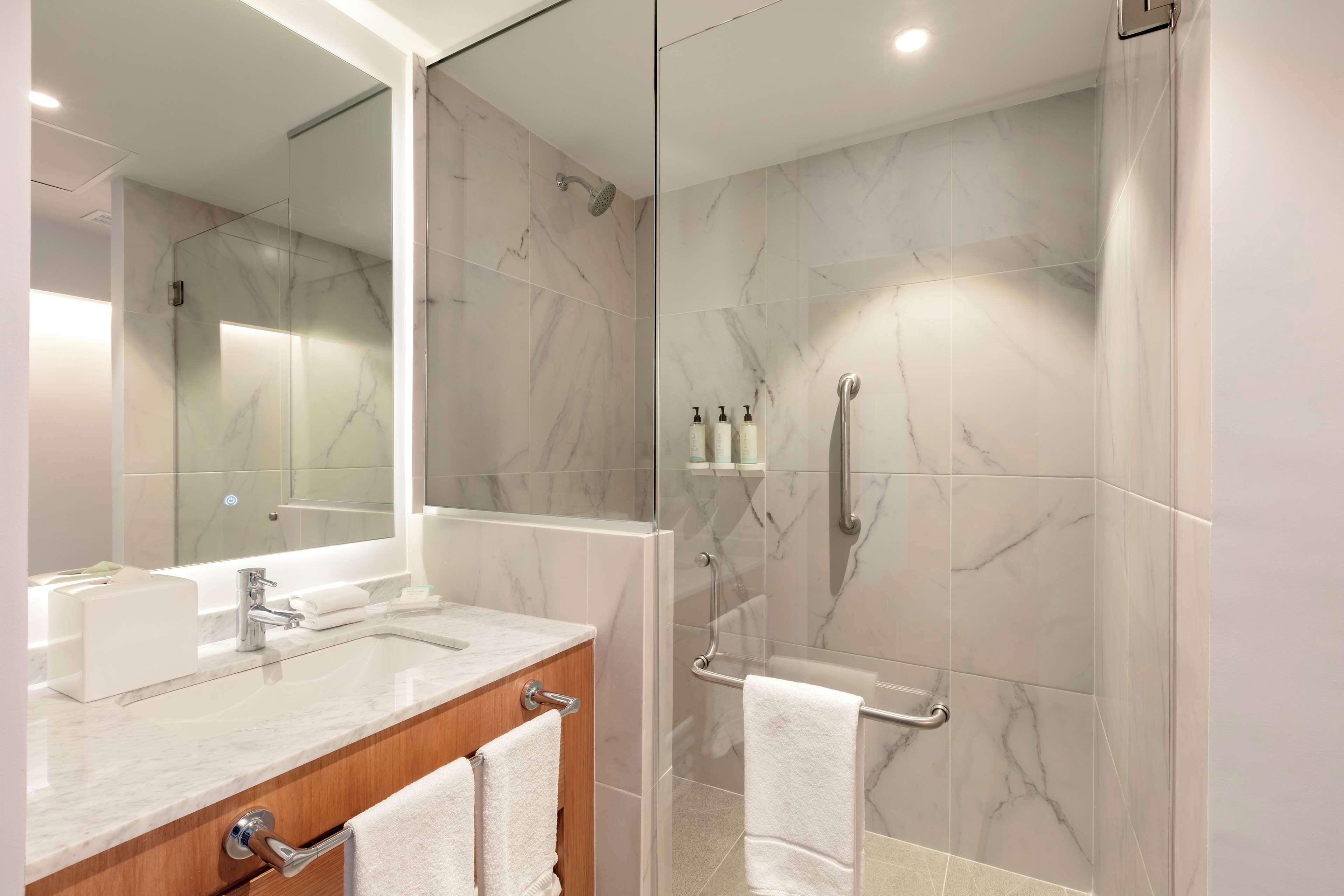 Un baño moderno con ducha revestida de mármol, zona de ducha con cerramiento de vidrio, tocador con lavabo y un espejo grande.