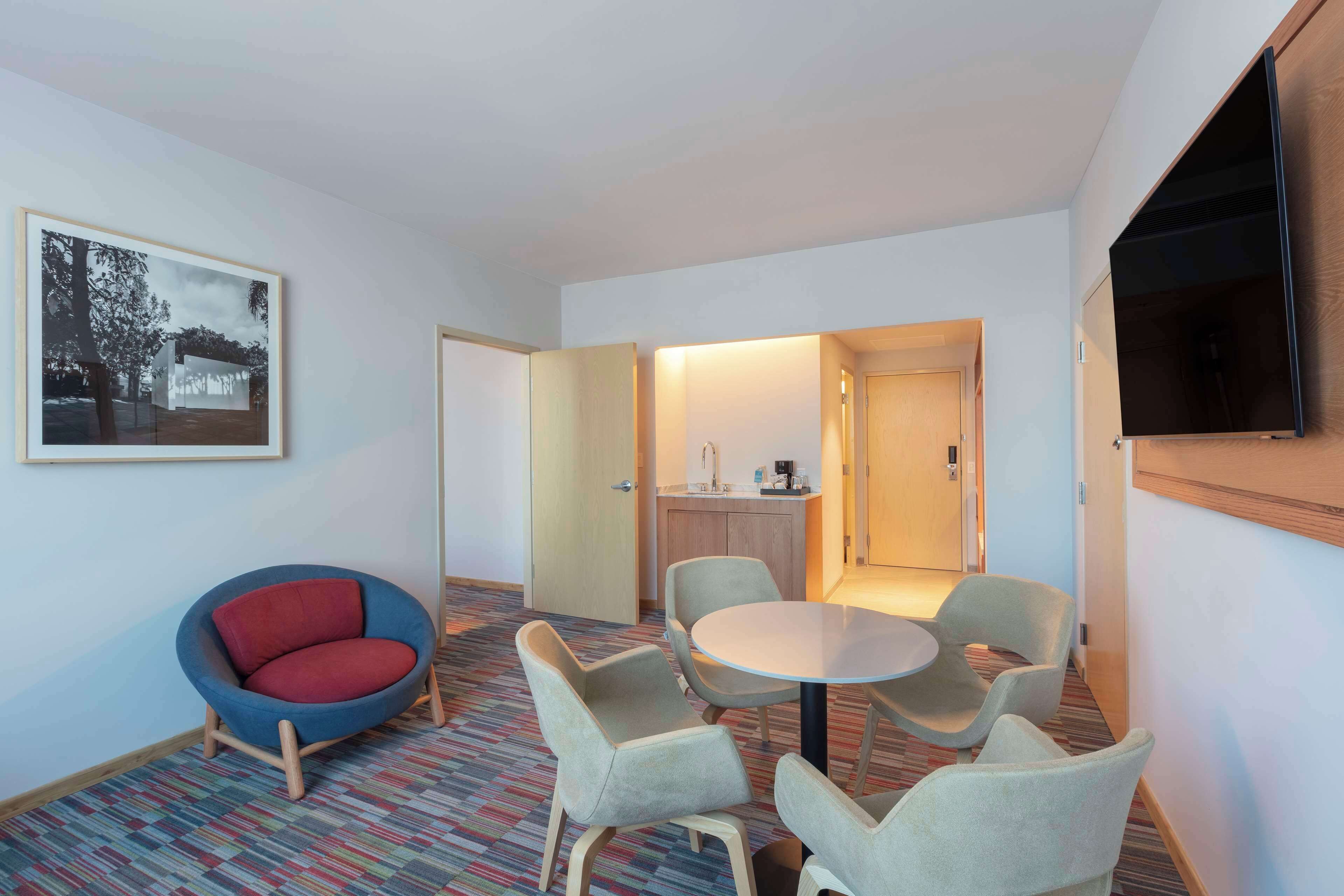 A hotel room with a round table, four chairs, a red armchair, a television, a framed picture on the wall, and a kitchenette in the background.
