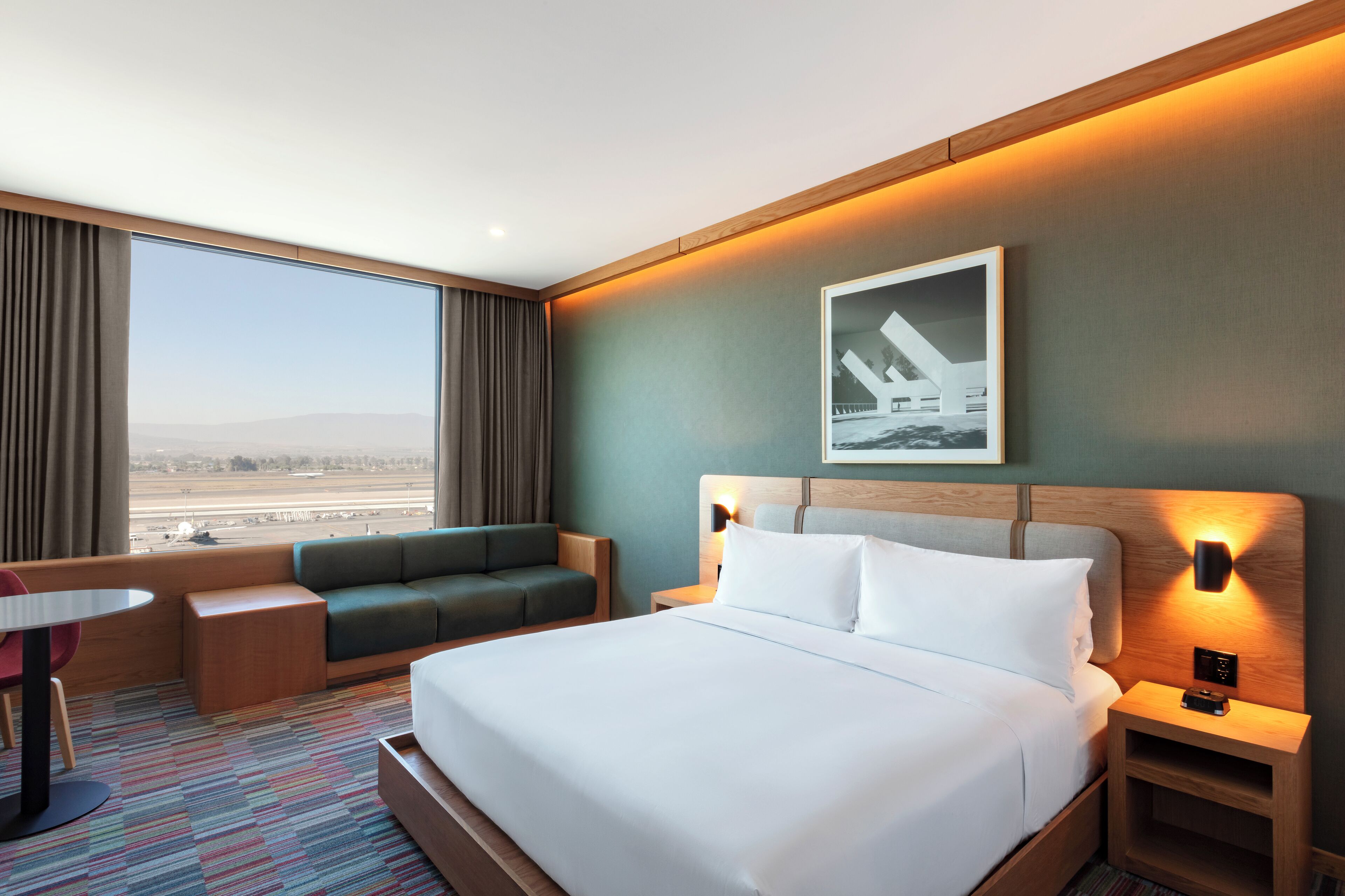 A hotel room with a large bed, a green sofa, a small table, and a view of the airport.
