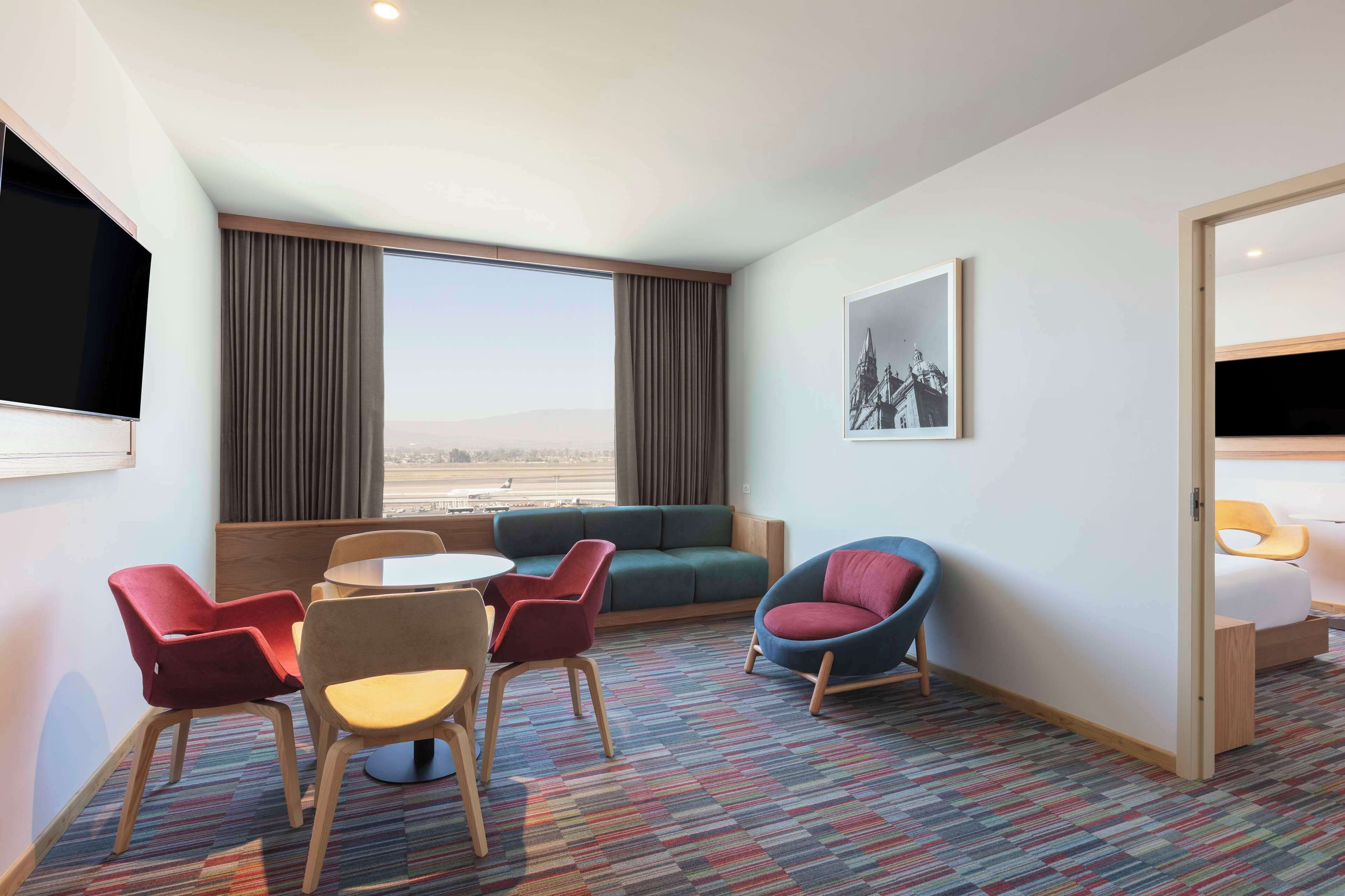 A hotel room with a large window, a sofa, a dining table with chairs, a TV, and a bed.