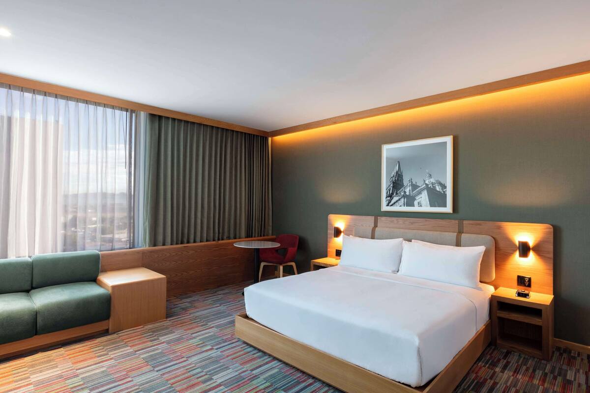 Une chambre d’hôtel avec un grand lit, un canapé vert, une petite table et une chaise. Il y a une fenêtre avec des rideaux, un tableau accroché au mur et des lampes de chevet.