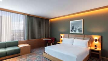 Habitación de hotel con una cama grande, un sofá verde, una mesita y una silla. Se aprecia una ventana con cortinas, un cuadro en la pared y lámparas de noche.