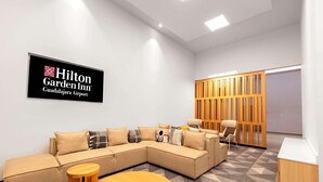 Un moderno y luminoso vestíbulo de hotel con un amplio sofá modular, una mesa de centro redonda de madera y una zona de estar con separador de madera.
