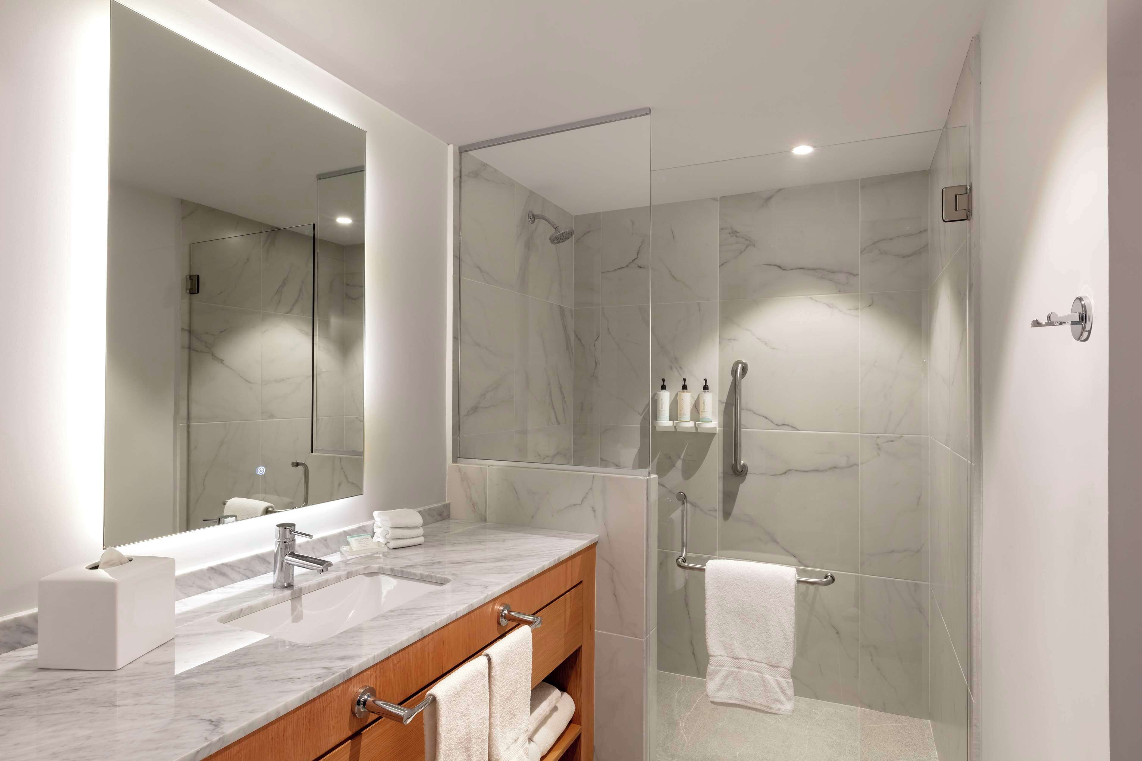 Un baño moderno con ducha envuelta en vidrio, paredes con azulejos de mármol, un espejo grande y un tocador con encimera de mármol.