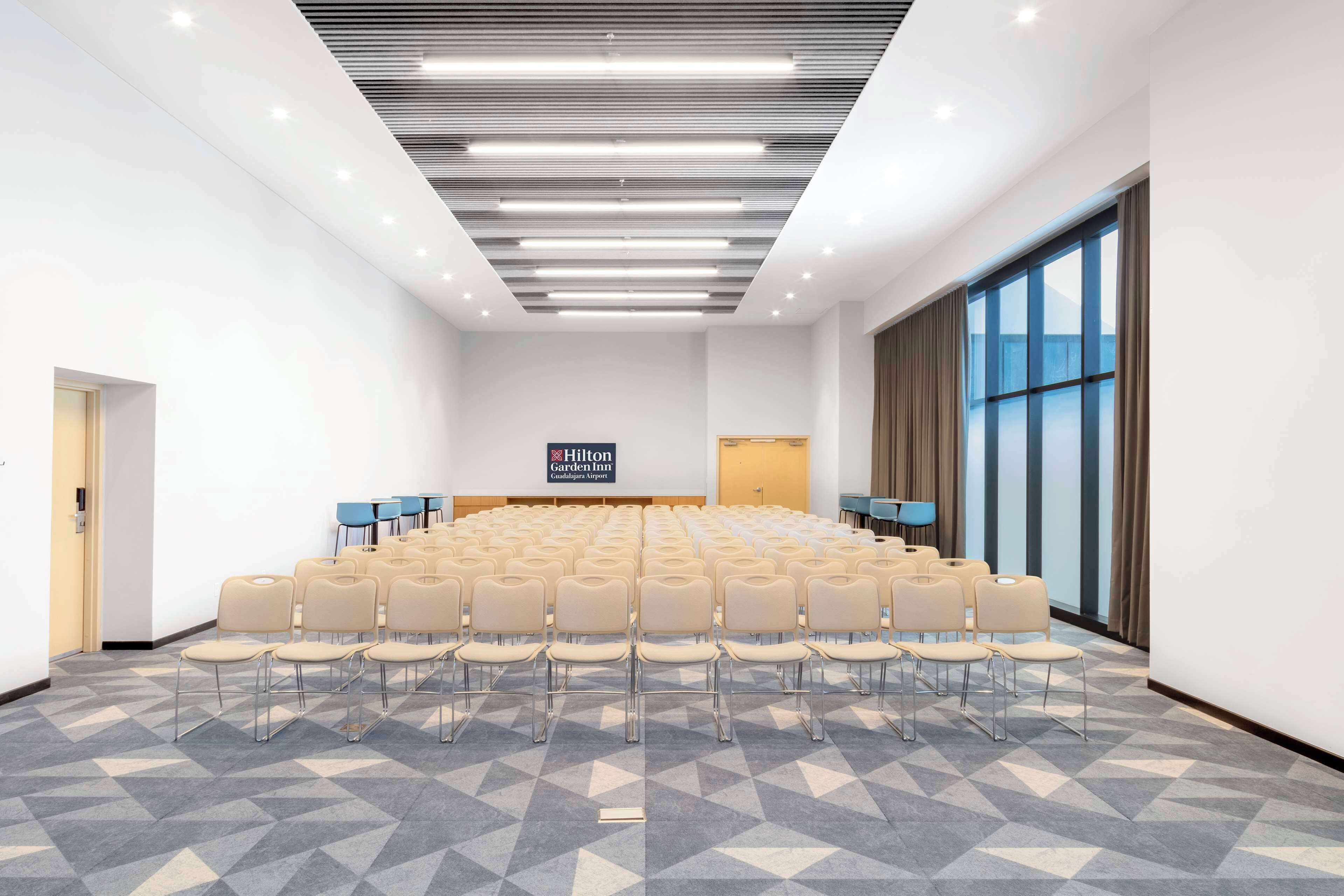 Un salón de conferencias con filas de sillas, un ventanal grande y un cartel de Hilton Garden Inn.