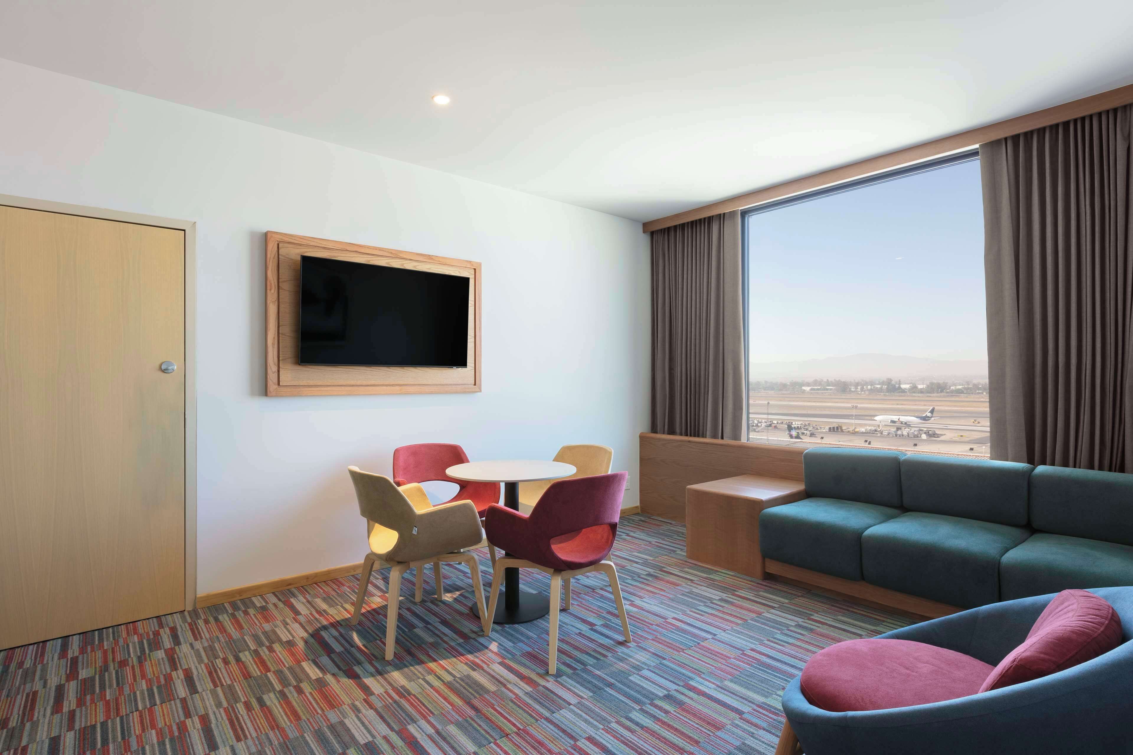 Una habitación de hotel con un ventanal grande, un televisor, un sofá y una pequeña mesa con sillas.