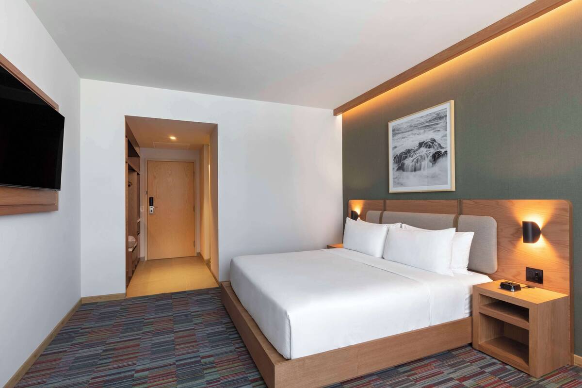 Une chambre d’hôtel avec un grand lit, une table de chevet avec une lampe, un téléviseur à écran plat fixé au mur et un tableau encadré au-dessus du lit.