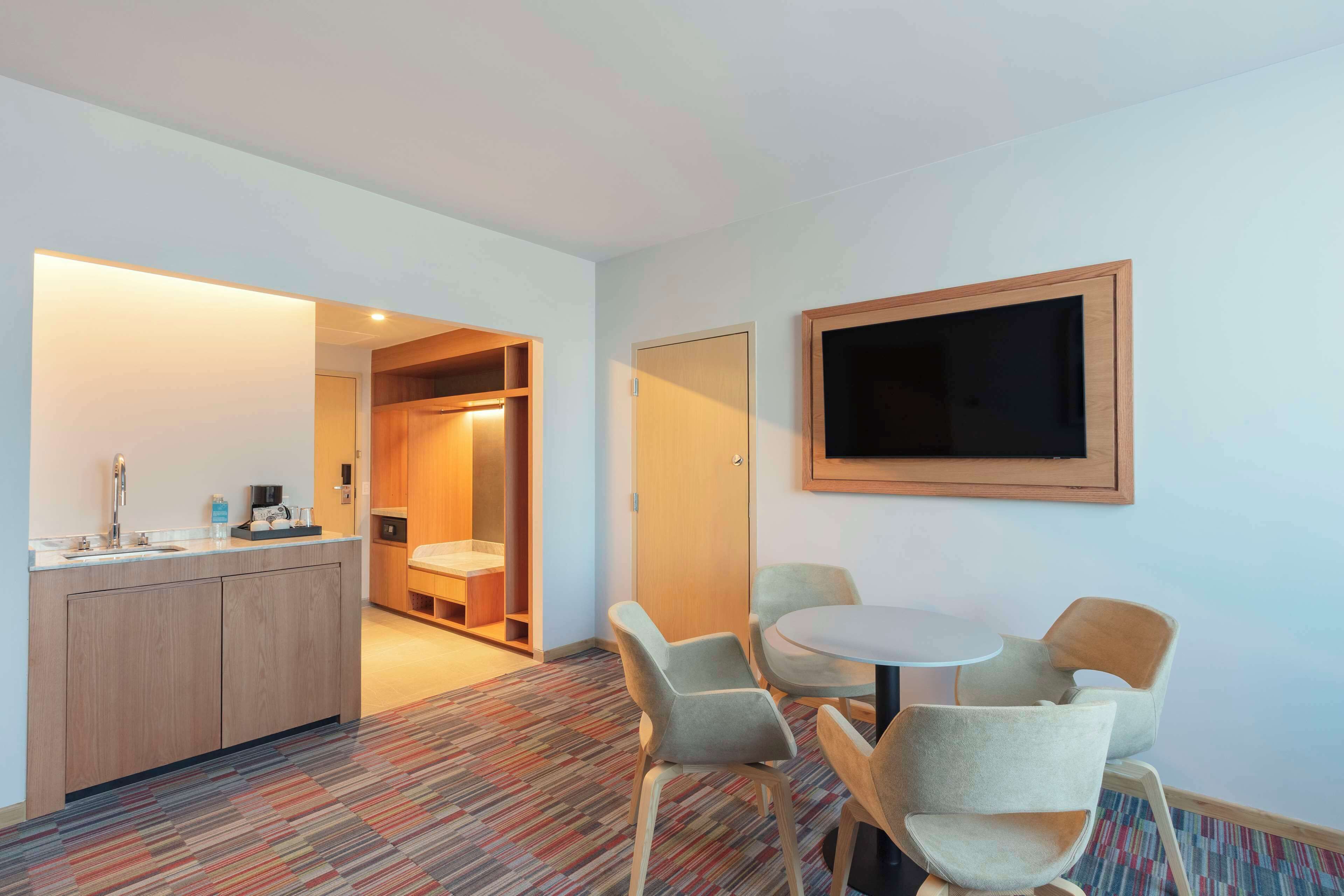 A hotel room with a kitchenette, a round dining table with four chairs, a flat-screen TV mounted on the wall, and a patterned carpet.