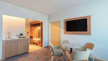 A hotel room with a kitchenette, a round dining table with four chairs, a flat-screen TV mounted on the wall, and a patterned carpet.
