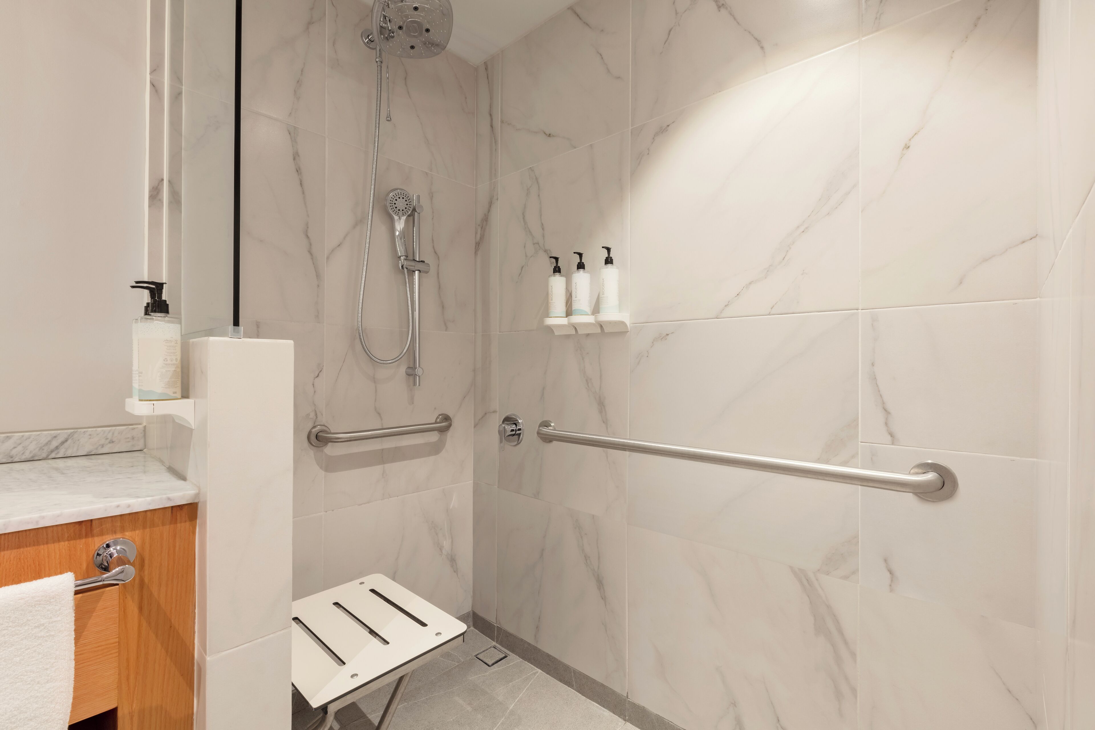A bathroom with marble walls, a shower with a handheld showerhead, a grab bar, and a small white bench.