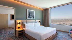 Una habitación de hotel con una cama grande, una mesita de noche, un cuadro en la pared, vistas al aeropuerto y una puerta.