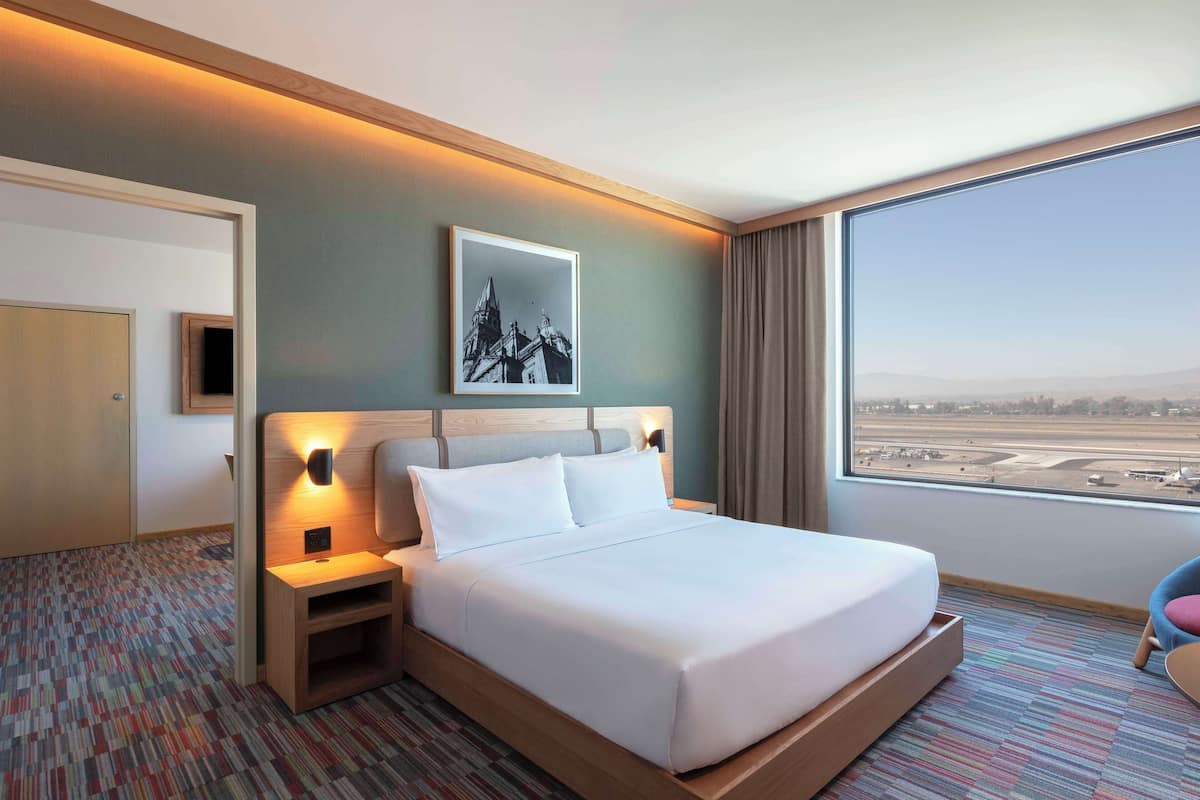 Une chambre d’hôtel avec un grand lit, une table de chevet, un tableau accroché au mur, une vue sur un aéroport et une porte.