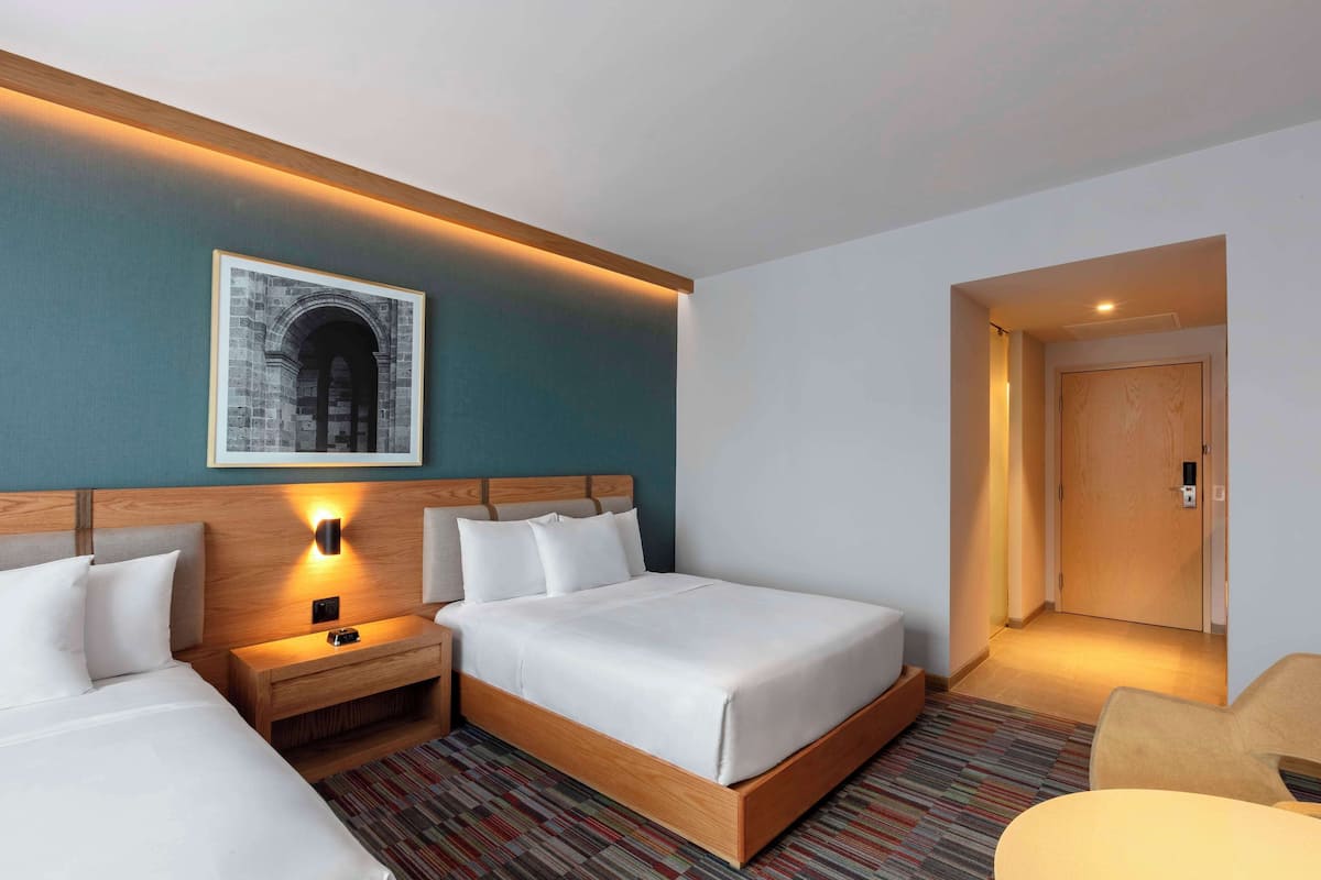 Une chambre d’hôtel avec un grand lit, une table de chevet avec une lampe, un tableau encadré au mur et une porte donnant sur une autre pièce.