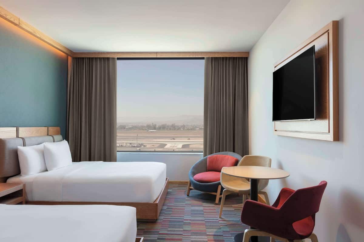 Une chambre d’hôtel avec un grand lit, un bureau, deux chaises, une télévision et une fenêtre donnant sur un aéroport.