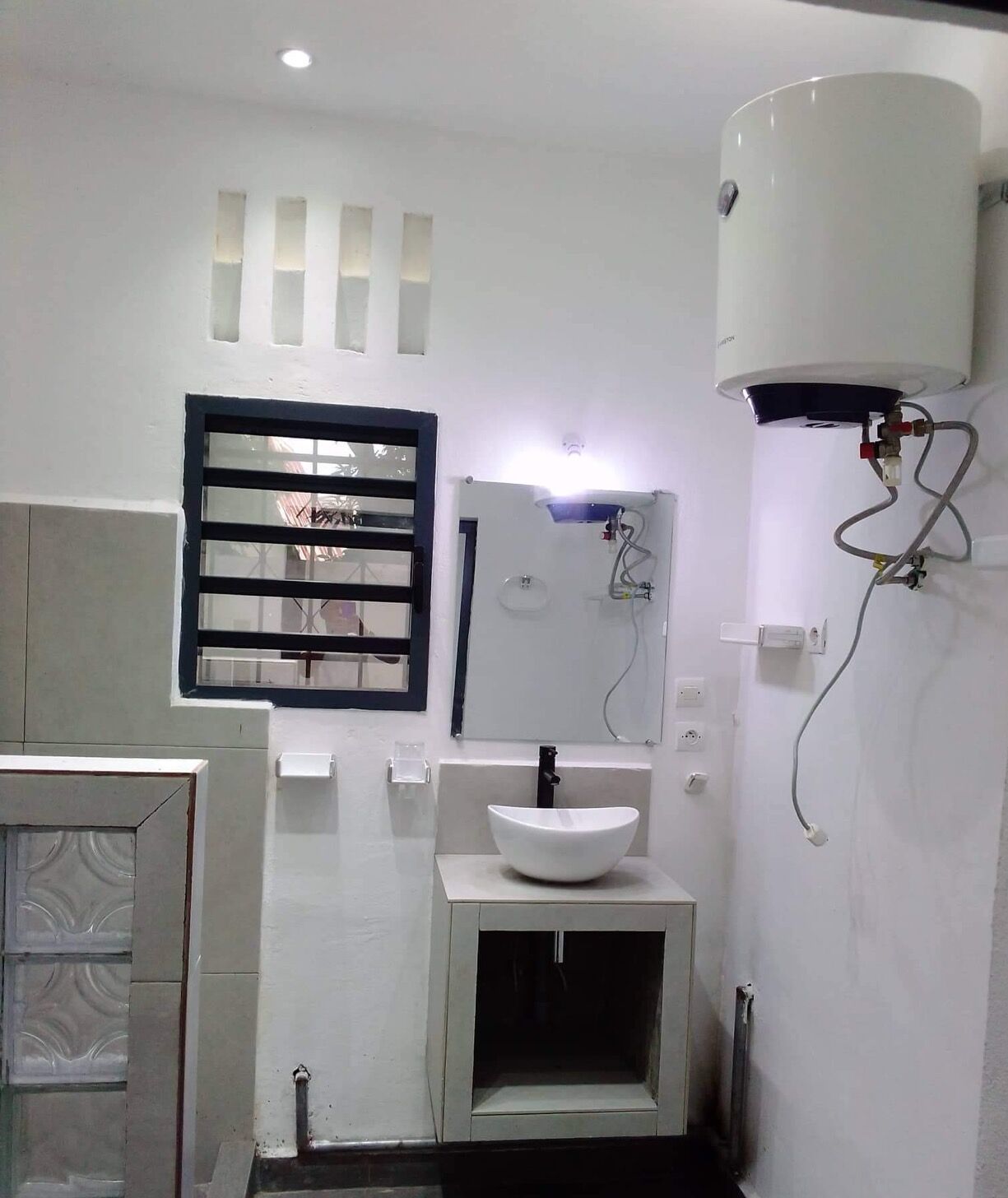 Une salle de bain avec un lavabo blanc, une fenêtre avec des stores horizontaux, un chauffe-eau et un miroir.