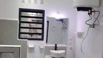 Une salle de bain avec un lavabo blanc, une fenêtre avec des stores horizontaux, un chauffe-eau et un miroir.