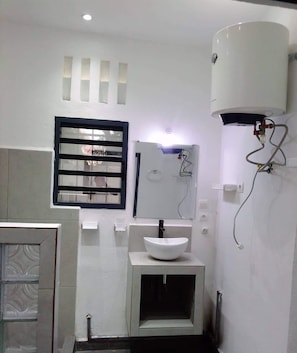 Un baño con un lavamanos blanco, una ventana con persianas horizontales, un calentador de agua y un espejo.