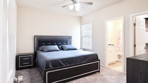 Une chambre à coucher comprenant un lit, des tables de chevet, un ventilateur de plafond et une porte donnant sur une salle de bain.