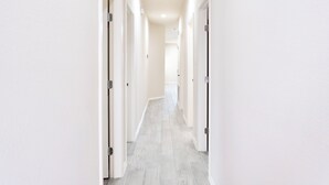 Un couloir aux murs blancs et au sol carrelé de couleur claire, qui mène à une pièce dont la porte se trouve au fond.