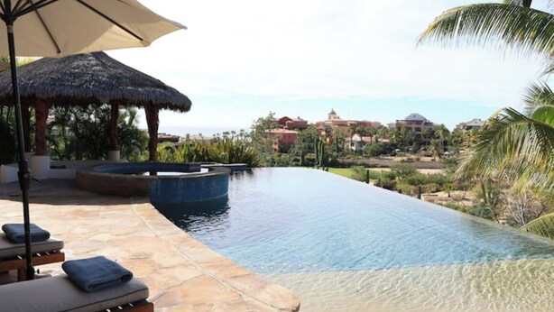 A swimming pool with a thatched umbrella, lounge chairs, and a view of a resort area. - Villa de Lujo Buena Vista 3 (Cabo San Lucas)