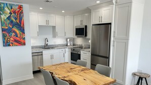 A modern kitchen with white cabinets, stainless steel appliances, and a wooden dining table. - Sea Glass at the Reef - Luxury King Beach Front Condo On The Beach (Colliers Bay)