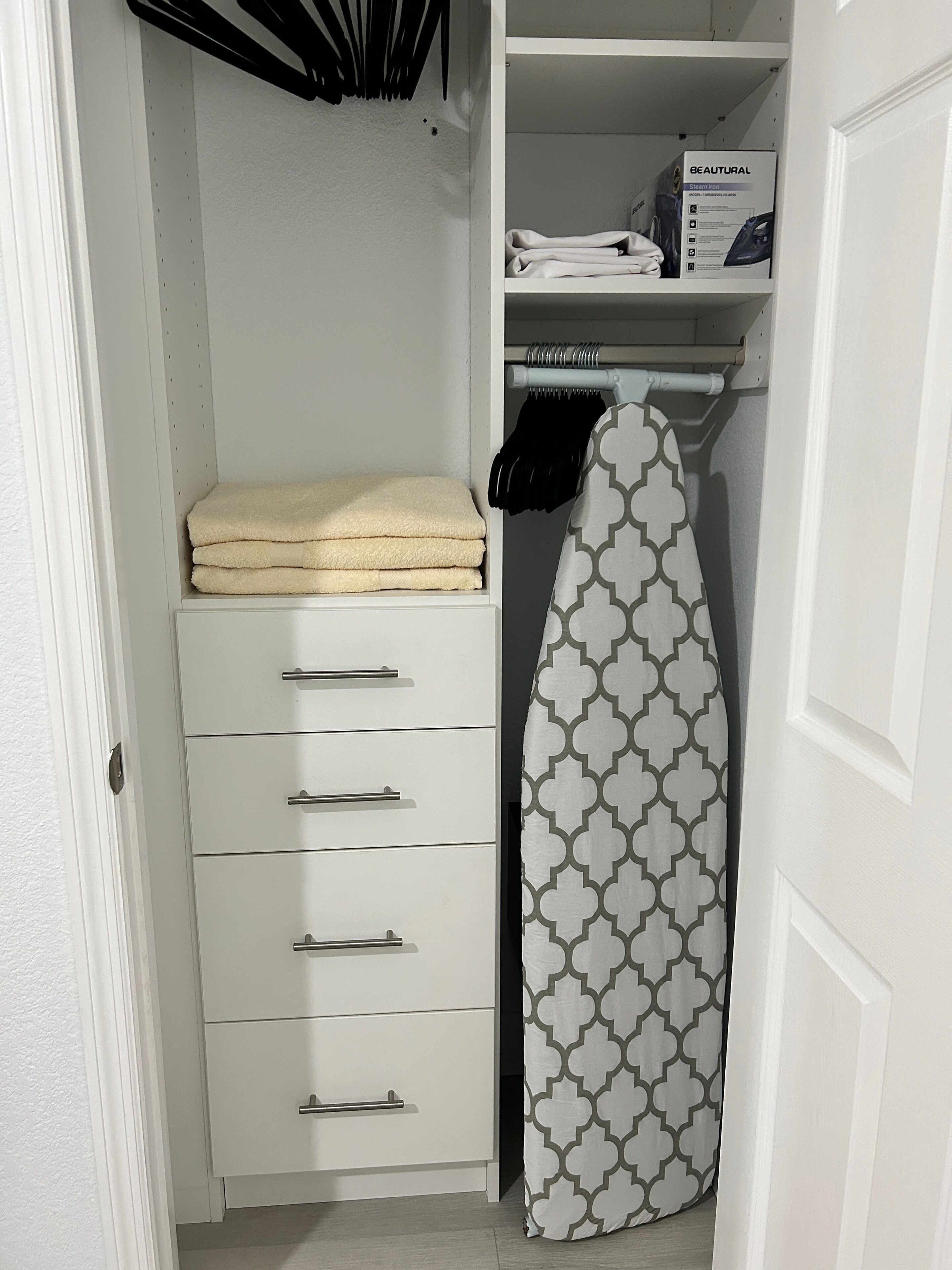 Un armario ordenado con un plancha colgando, toallas dobladas y una caja etiquetada 'BEAUTURAL Steam Iron'.