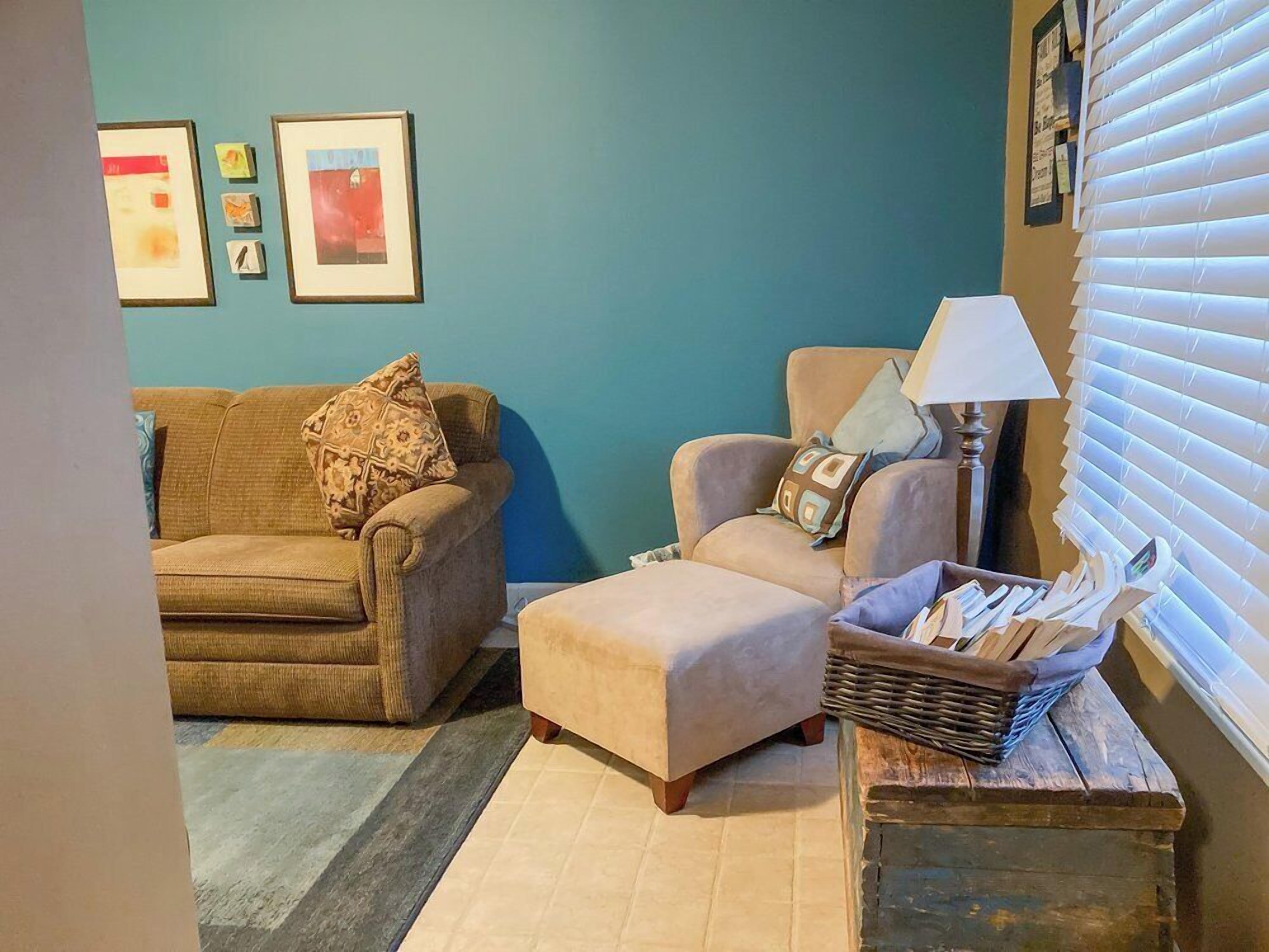 A living room with a brown sofa, a beige armchair, and a small table with a lamp and a basket of magazines.